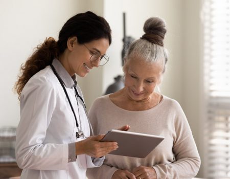 Happy female doctor and senior patient look at tablet screen discuss treatment or therapy on gadget. Smiling woman nurse and mature client use pad device discussing results in modern hospital.