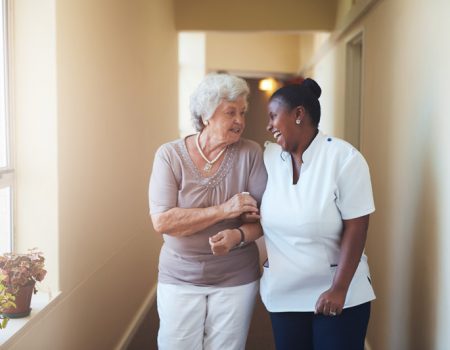 Portrait of happy female caregiver and senior woman walking together at home. Professional caregiver taking care of elderly woman.