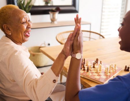 Black people, nurse and high five in elderly care for chess, fun or social activity or game together at home. African medical professional touching hands with senior female person with a disability.
