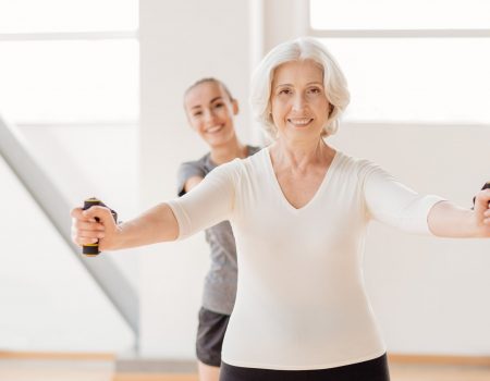 Involved in the workout. Hard working pleasant elderly woman standing in front of her coach and exercising with a resistance band while having fitness classes