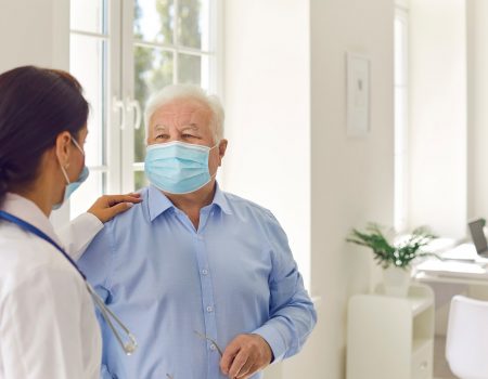 Young professional woman doctor touching shoulder of mature senior man patient in mask after visit in modern light clinic interior. Visiting doctor durinf pandemic concept