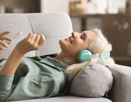 Happy blonde old woman in big headphones listening to music with closed eyes from online playlist, using online media service, application on smartphone, lying on home couch